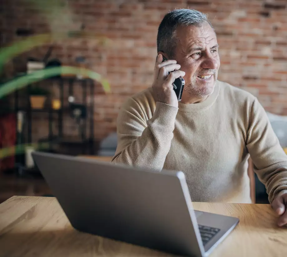Un homme cinquantenaire riant au téléphone. Il est assis à un bureau sur lequel il y a un ordinateur portable.