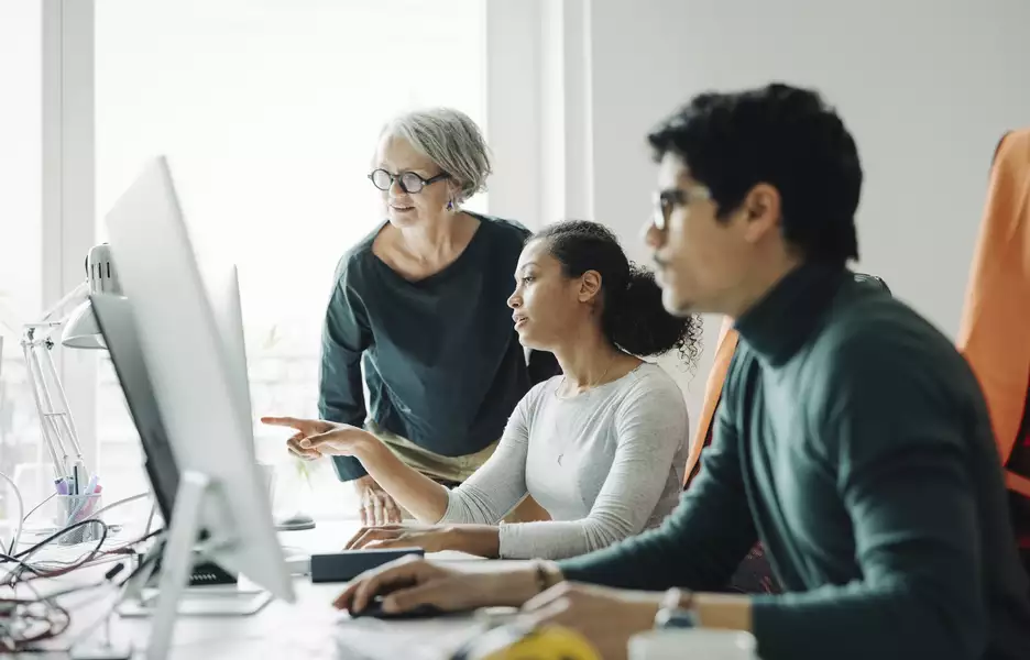 2 personnes qui travaillent assis à leur bureau et 1 personne qui vient regarder l'écran